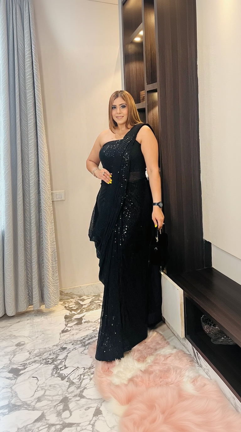 A woman wearing a black sequin saree gown with a floral design, featuring a one-shoulder with strap on the lift side, standing in a luxurious interior.