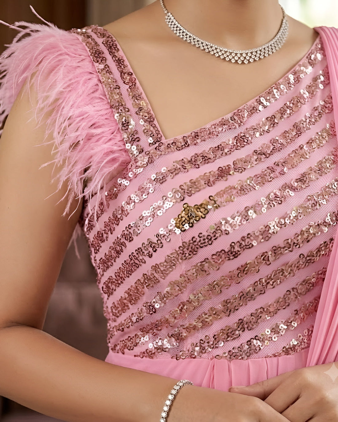 A close-up of a woman wearing a pink embellished blouse with sequined diagonal stripes, feathered shoulder detail, and a draped pink fabric, paired with a silver necklace and bracelet.