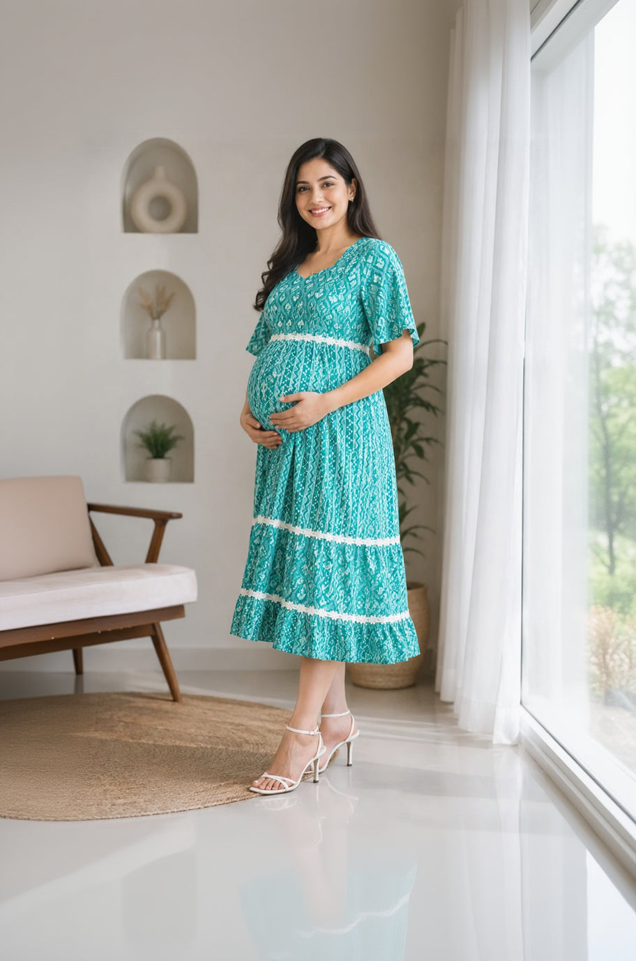 Pregnant woman in a teal dress with white trim standing in a room with plants and a chair.