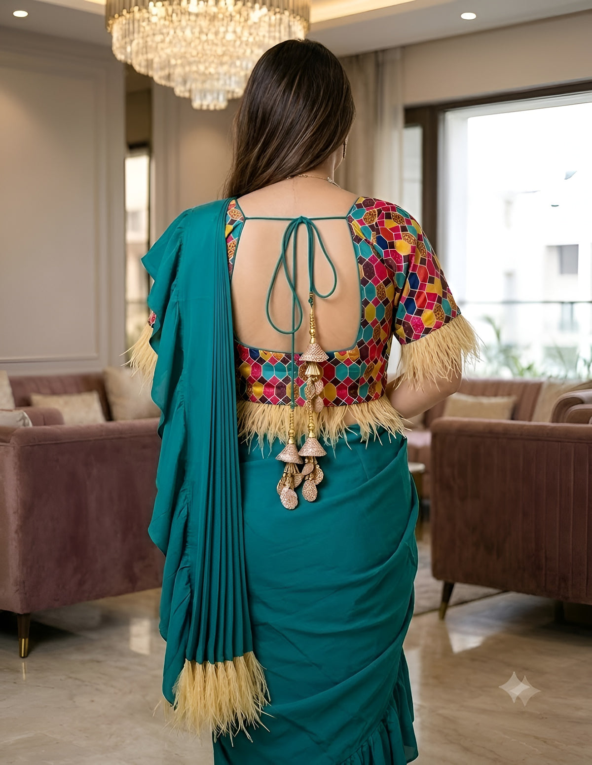 A woman shown from behind wearing a teal green saree with a highly colorful, patterned blouse featuring yellow fringe and hanging tassel ornaments, standing in a modern living room.