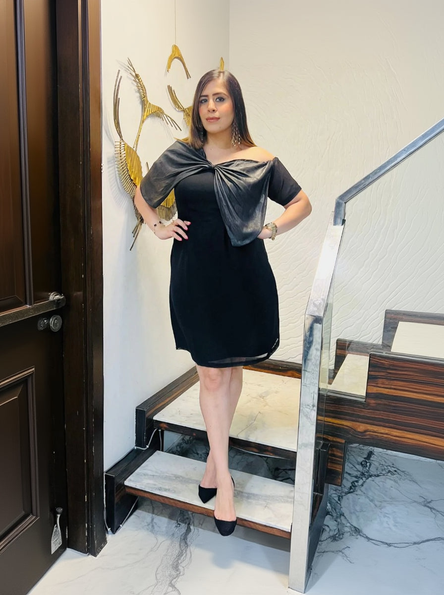 A woman standing on a staircase wearing a black dress with a bow detail at the neck, posing with one hand on her hip.