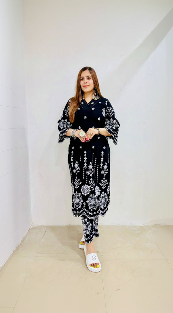 A woman standing against a white background, wearing a black embroidered cotton suit with a V-neckline, 3/4 sleeves, and side slits, paired with pants.