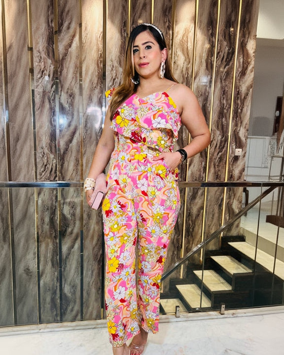 A woman wearing a pink floral jumpsuit with a one shoulder neckline and strappy detail on the other shoulder, standing in front of a wooden backdrop.