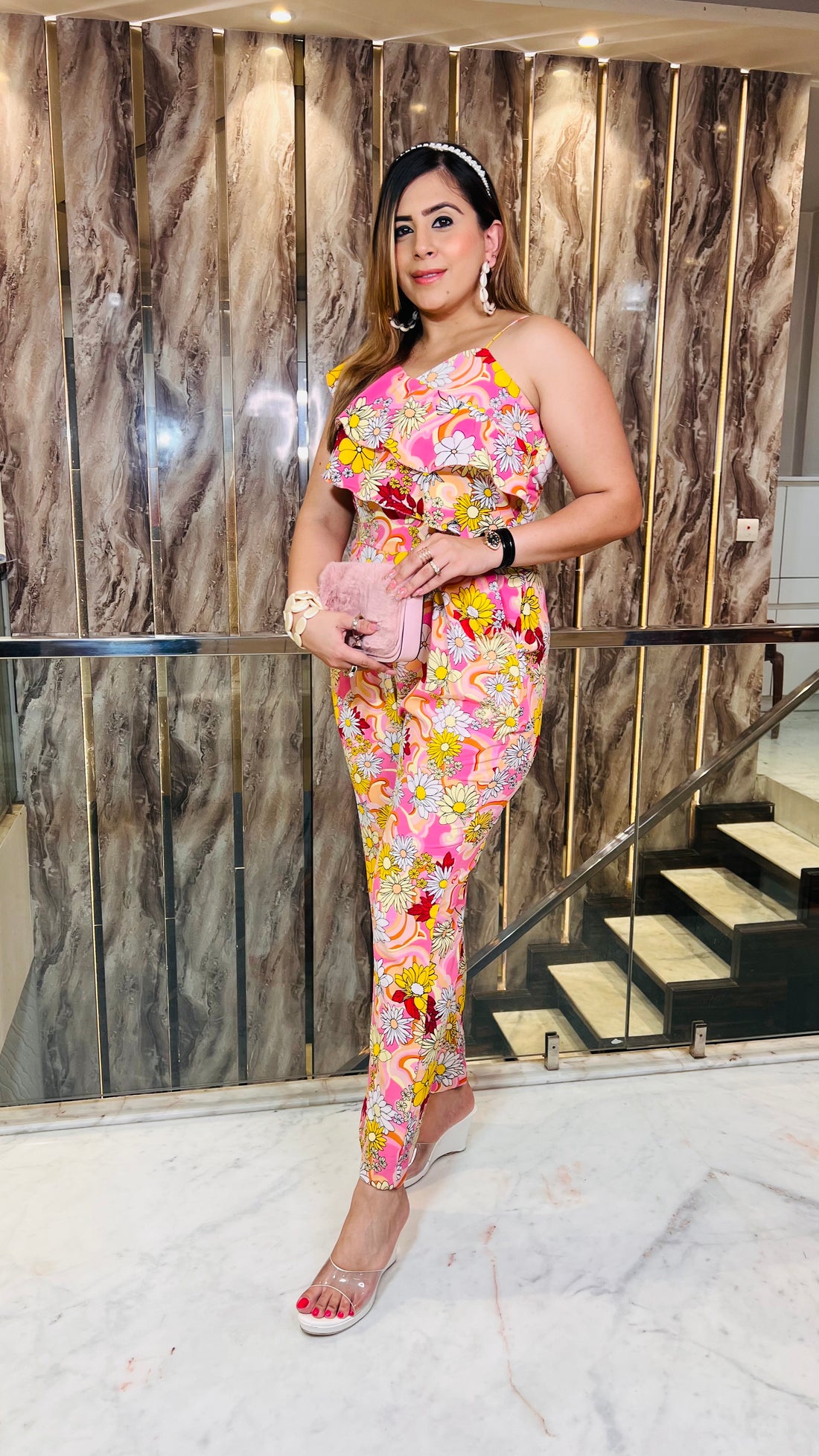 A woman wearing a pink floral jumpsuit with a one shoulder neckline and strappy detail on the other shoulder, standing in front of a wooden backdrop.
