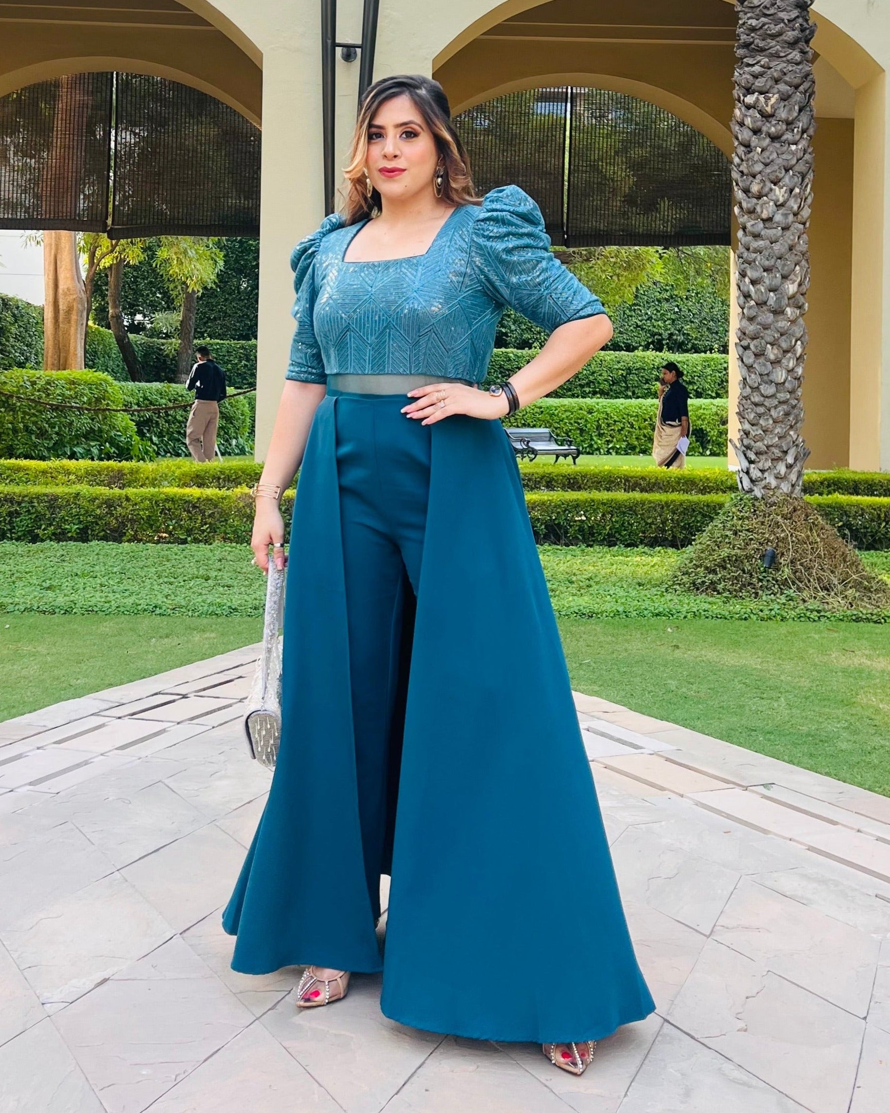 A woman standing outdoors wearing a teal blue party jumpsuit with elbow-length puffy sleeves, a square neckline, and cross pockets. She is also wearing high heels and posing in front of a building with palm trees in the background.