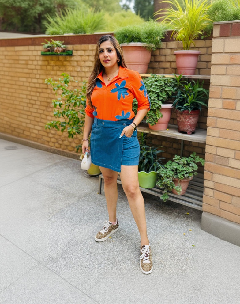 Woman in an orange shirt with blue patterns and denim shorts standing in front of a plant-filled outdoor area.