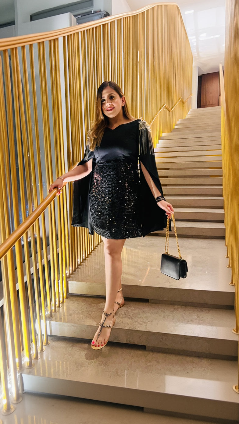 A woman standing on a staircase wearing a black sequin dress with off-shoulder sleeves and cap sleeves, holding a black purse and wearing open-toe heels.