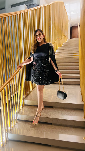 A woman standing on a staircase wearing a black sequin dress with off-shoulder sleeves and cap sleeves, holding a black purse and wearing open-toe heels.