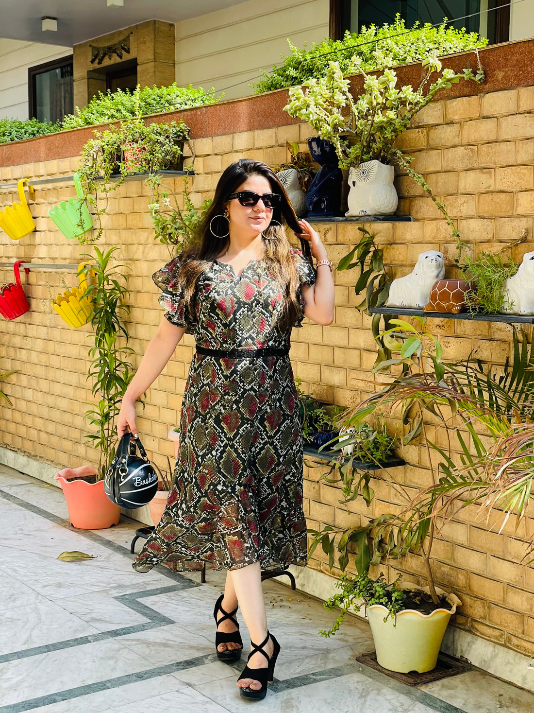 A woman standing on a balcony wearing a black snakeskin print midi dress with round neckline and frill detailing on the shoulders, paired with black sandals and sunglasses.