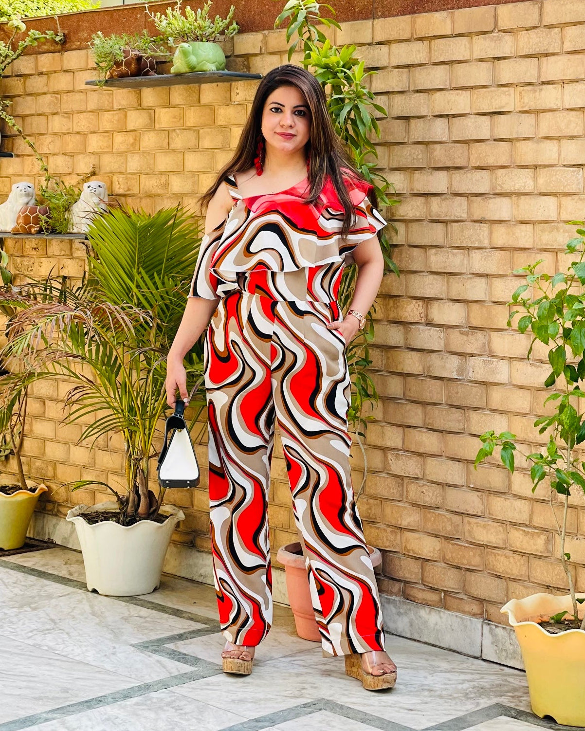 A person wearing a red wiggle jumpsuit with a one shoulder neck style, standing near a brick wall with plants.