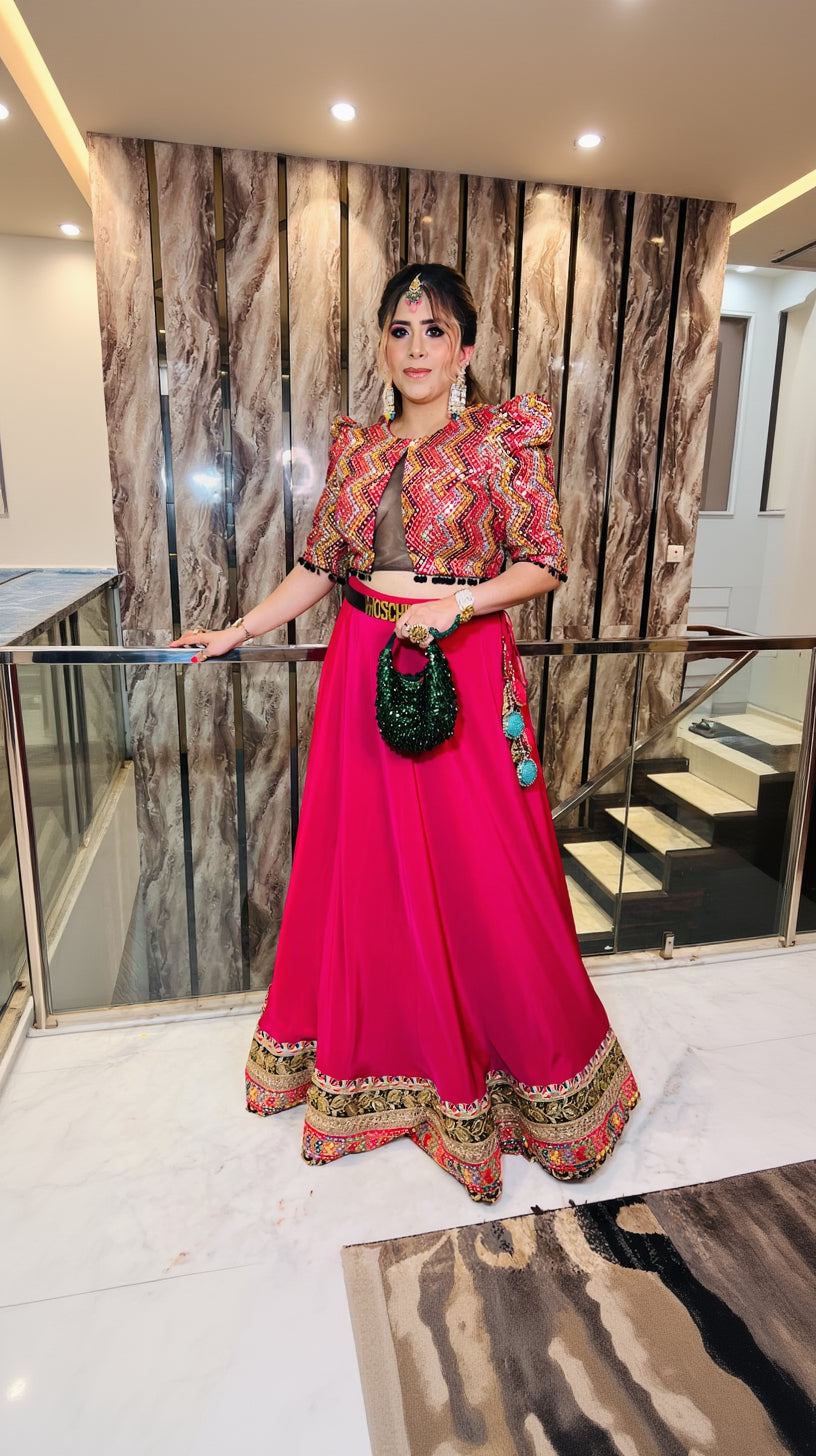 A woman wearing a pink mirror work lehenga with geometric patterns and black fringes lace on the blouse, paired with a belt and carrying a black purse.