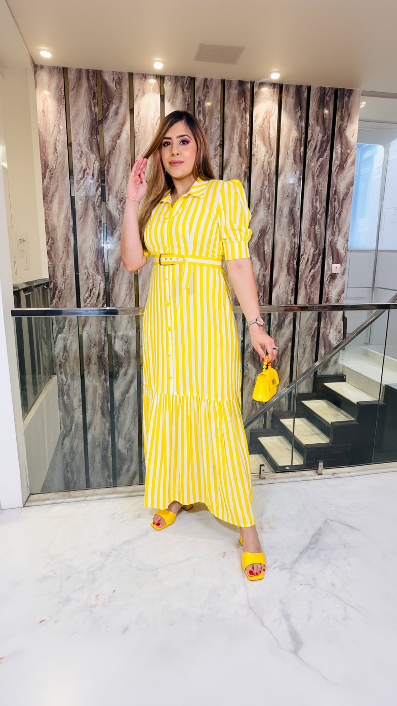 A woman wearing a yellow maxi dress with horizontal stripes, featuring a shirt collar and elbow length puffy sleeves, standing in a luxurious interior.
