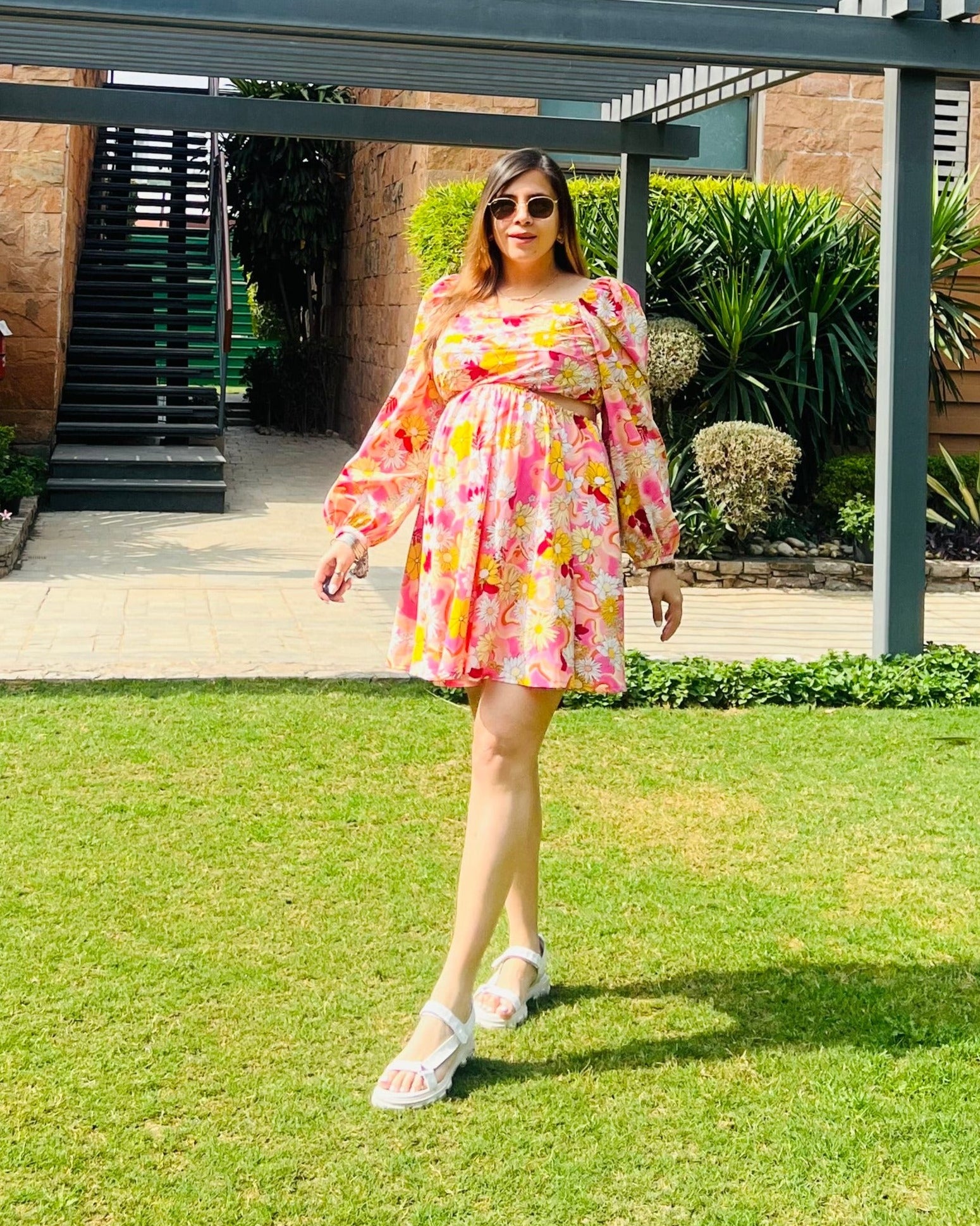 A woman standing on grass wearing a short-sleeved, pink, floral dress with a sweetheart neckline, elastic on the shoulders, and gathered fabric at the mid-section.