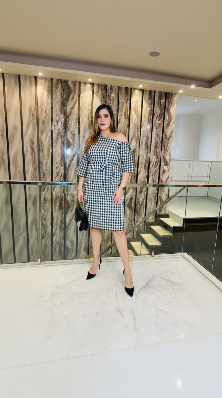 A woman wearing a black and white checkered off-shoulder dress with 3/4th sleeve on the right side and an elasticated sleeve on the left, paired with a belt of the same fabric.