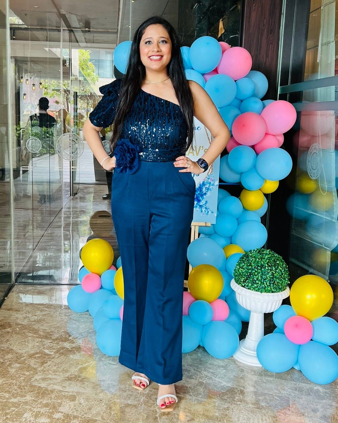 A person wearing a navy blue sequin jumpsuit with a flower belt standing in front of a wall with colorful balloons.