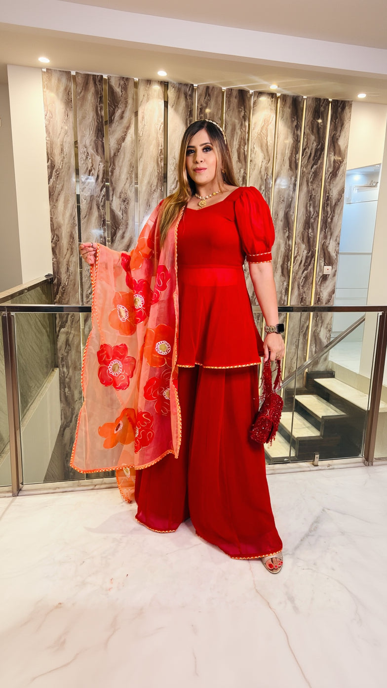 A woman wearing a red handpainted sharara suit with floral details and a sweetheart neckline top, holding a red flower, standing in a luxurious interior.