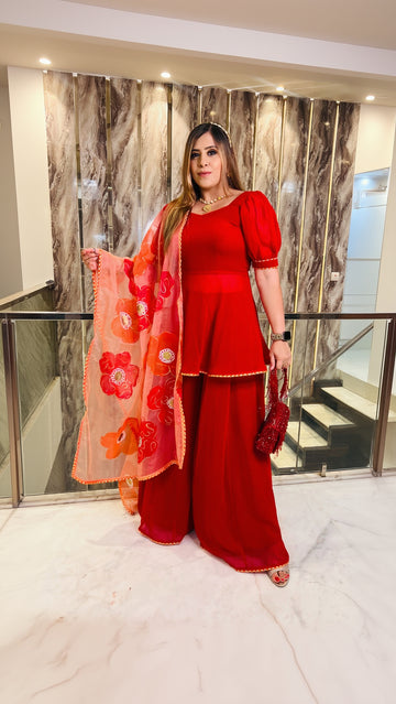 A woman wearing a red handpainted sharara suit with floral details and a sweetheart neckline top, holding a red flower, standing in a luxurious interior.