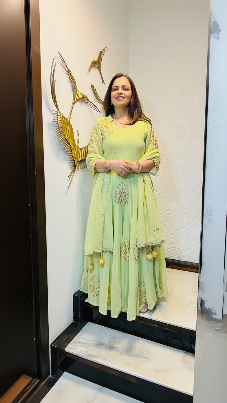 A woman standing and smiling, wearing a pastel green handwork anarkali with embroidery and a net dupatta.