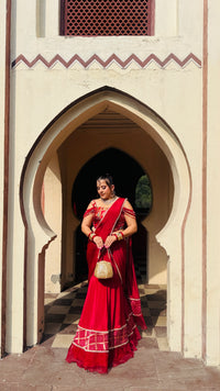 A woman wearing a vibrant red traditional Indian sari with gold embroidery stands in a stone archway doorway, holding a small woven bag.