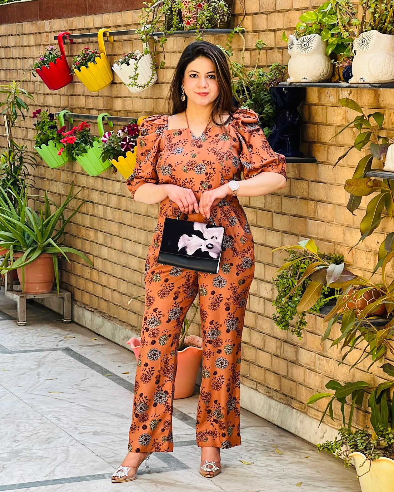 A woman standing in front of a wall with plants, wearing a peach jumpsuit with a square neck and puffed sleeves, with the fabric of the jumpsuit visible.