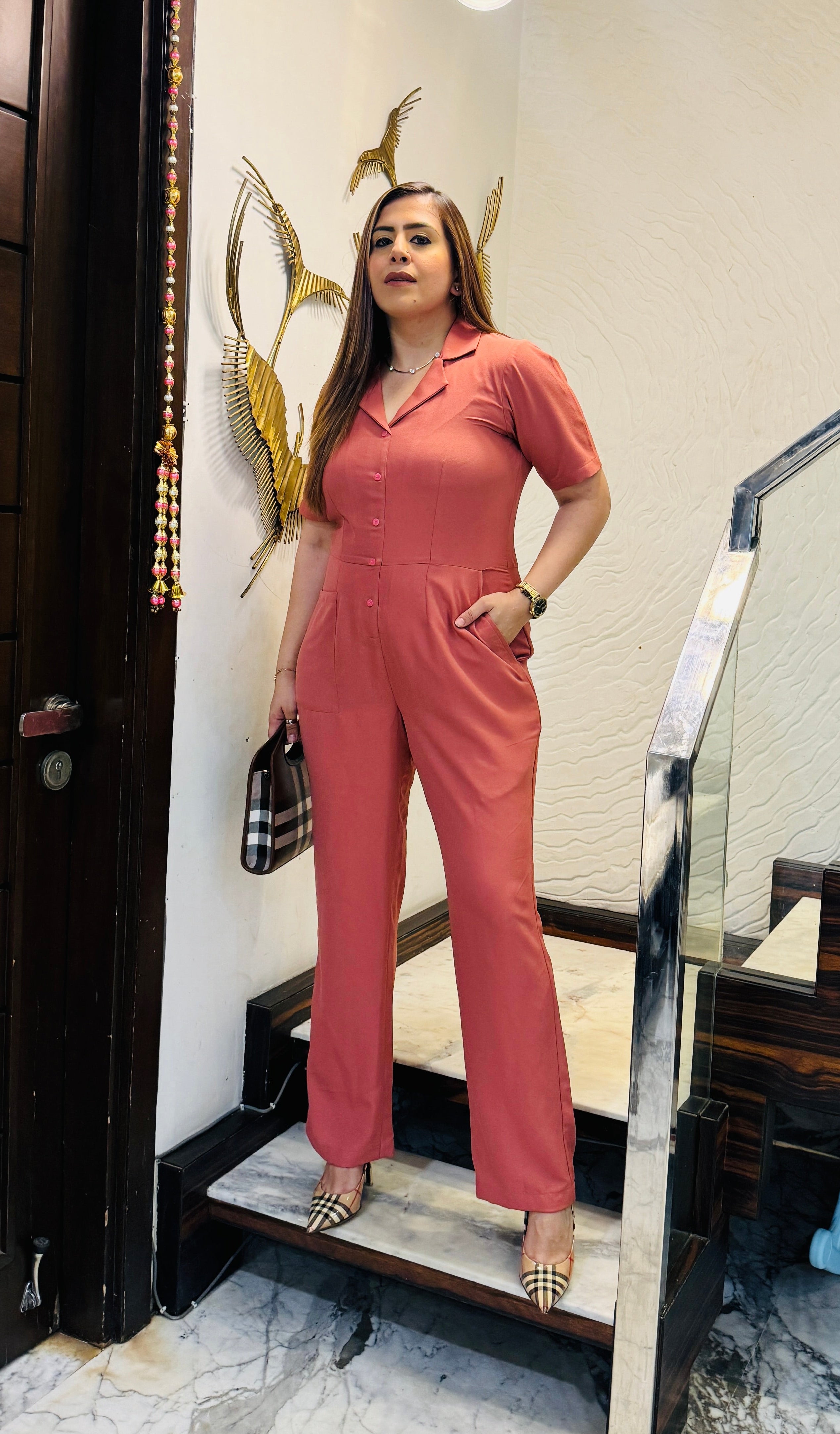 A woman standing on a staircase wearing a peach army jumpsuit with a collared neckline, front buttons, and pockets.
