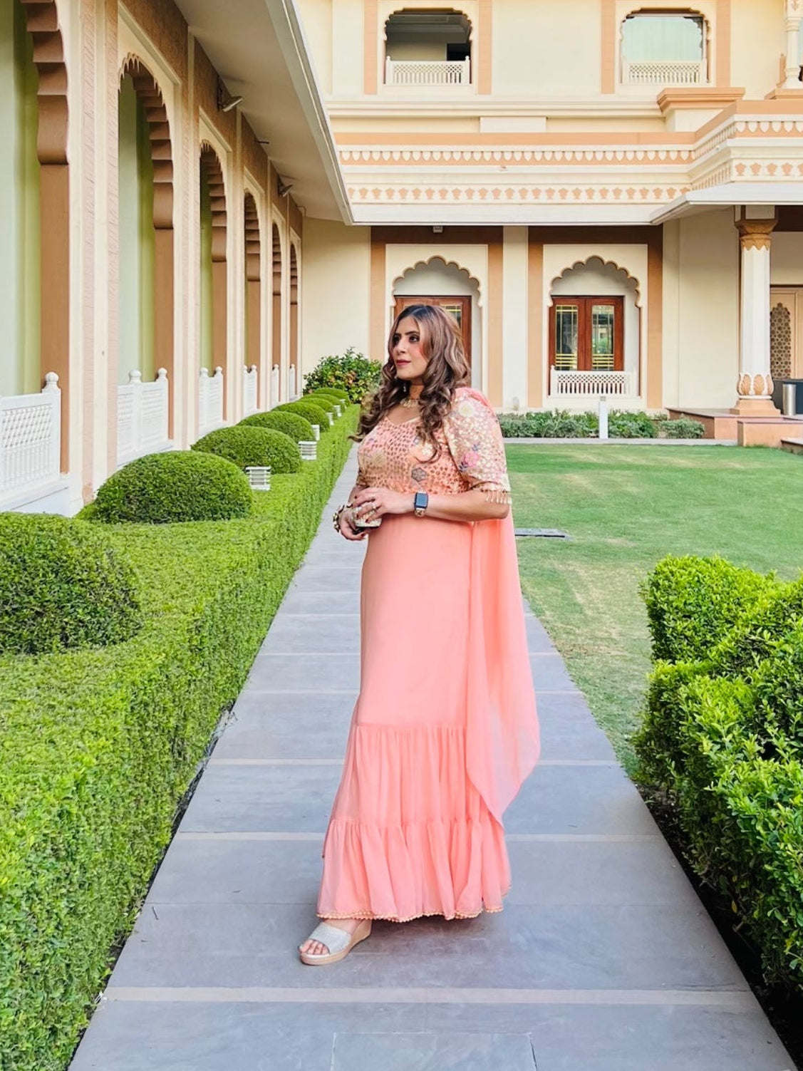 A person wearing a peach-colored traditional ethnic Indian dress with embroidered details, standing in a garden with archways in the background.