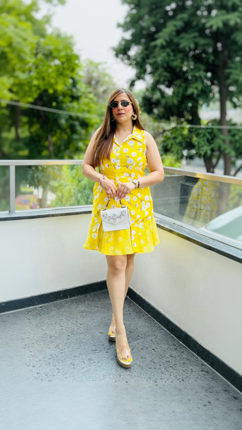 A woman wearing a sleeveless, floral print, yellow dress with ruffle details and a flared hem. She has accessorized with gold hoop earrings, sunglasses, and a small, white, jewel-encrusted purse.