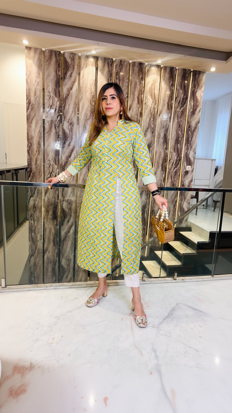 A woman wearing a pastel green, chevron patterned kurti with elbow length sleeves and a half neck cut, standing indoors with one hand on a railing.