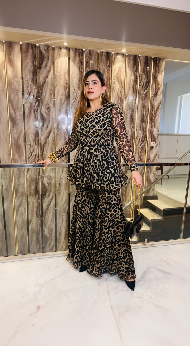 A woman wearing a traditional brown garara suit with a leopard print pattern, featuring a round neckline and full sleeves, standing in a luxurious setting.