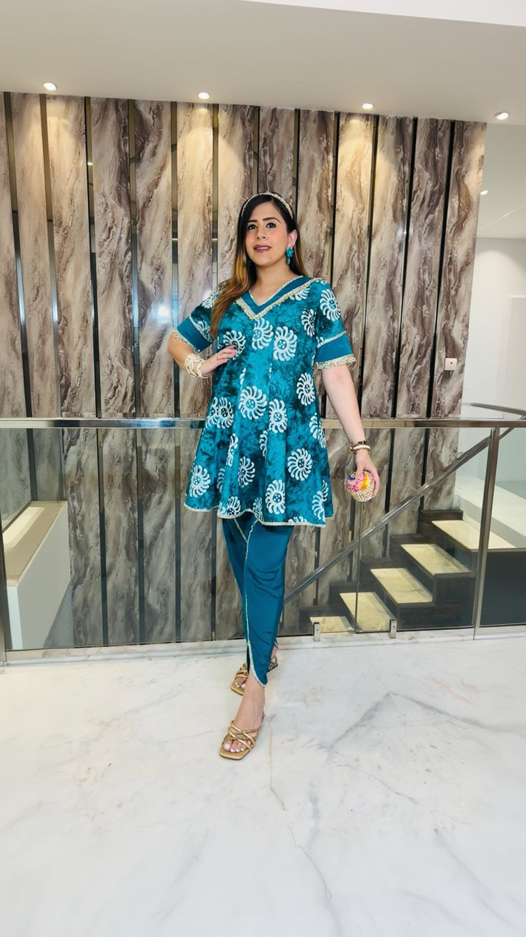 A woman wearing a teal blue cotton batik dhoti suit with a peplum top and pants, featuring a V-neck style and lace detailing on the sleeves.