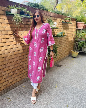 Woman in a pink floral dress standing outdoors near a wooden wall with plants.