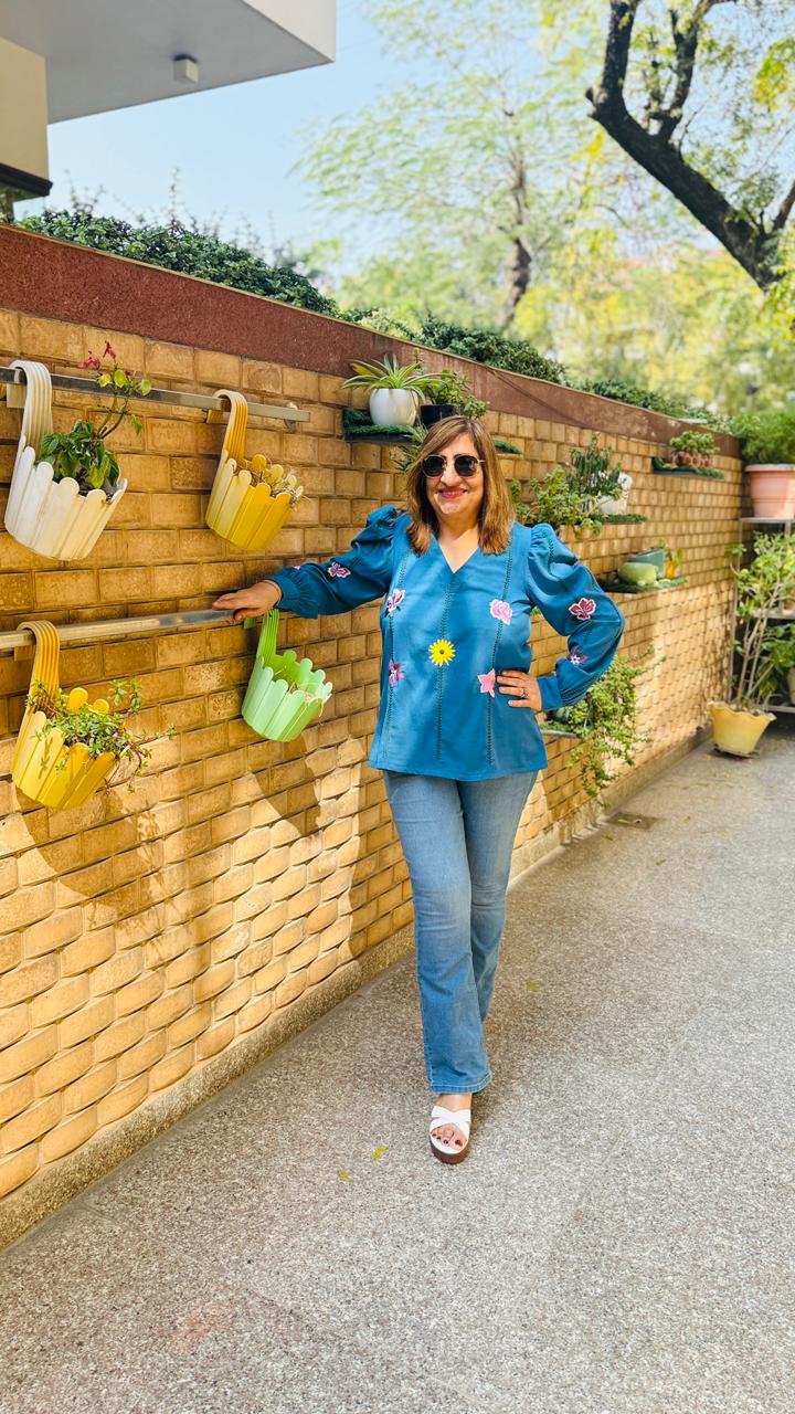 Woman in a blue jacket standing next to a wall with hanging plants on a sunny day.