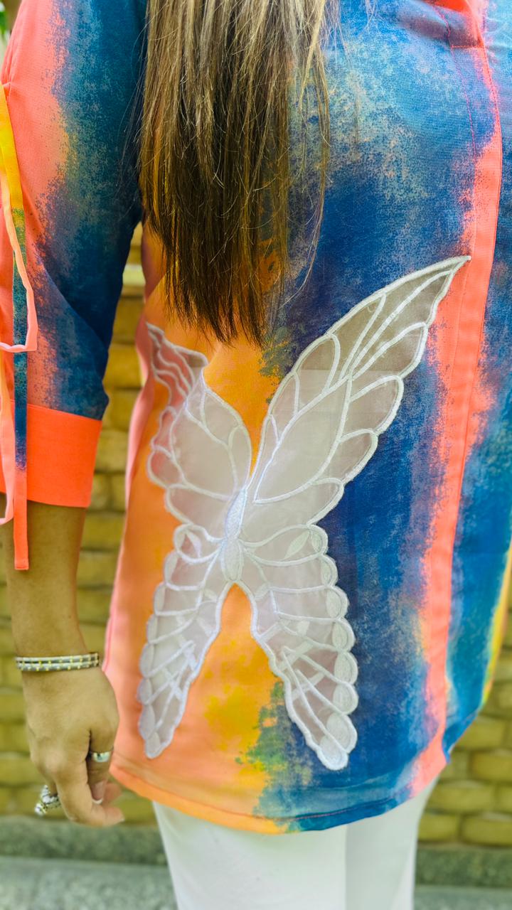 Woman in a colorful shirt and white pants standing in front of a wooden wall with plants.
