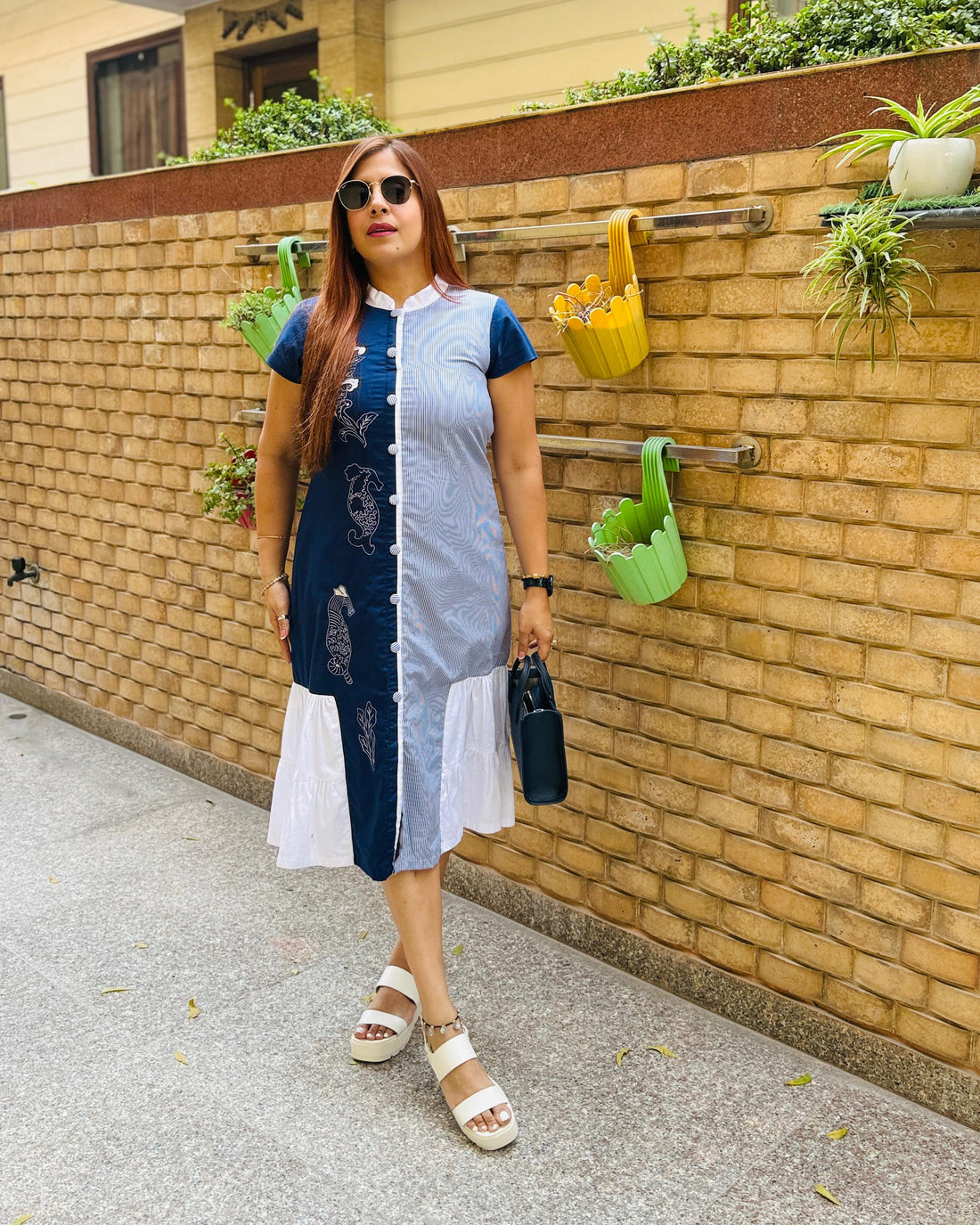 Woman standing in front of a brick wall with plants, wearing a blue and white dress.