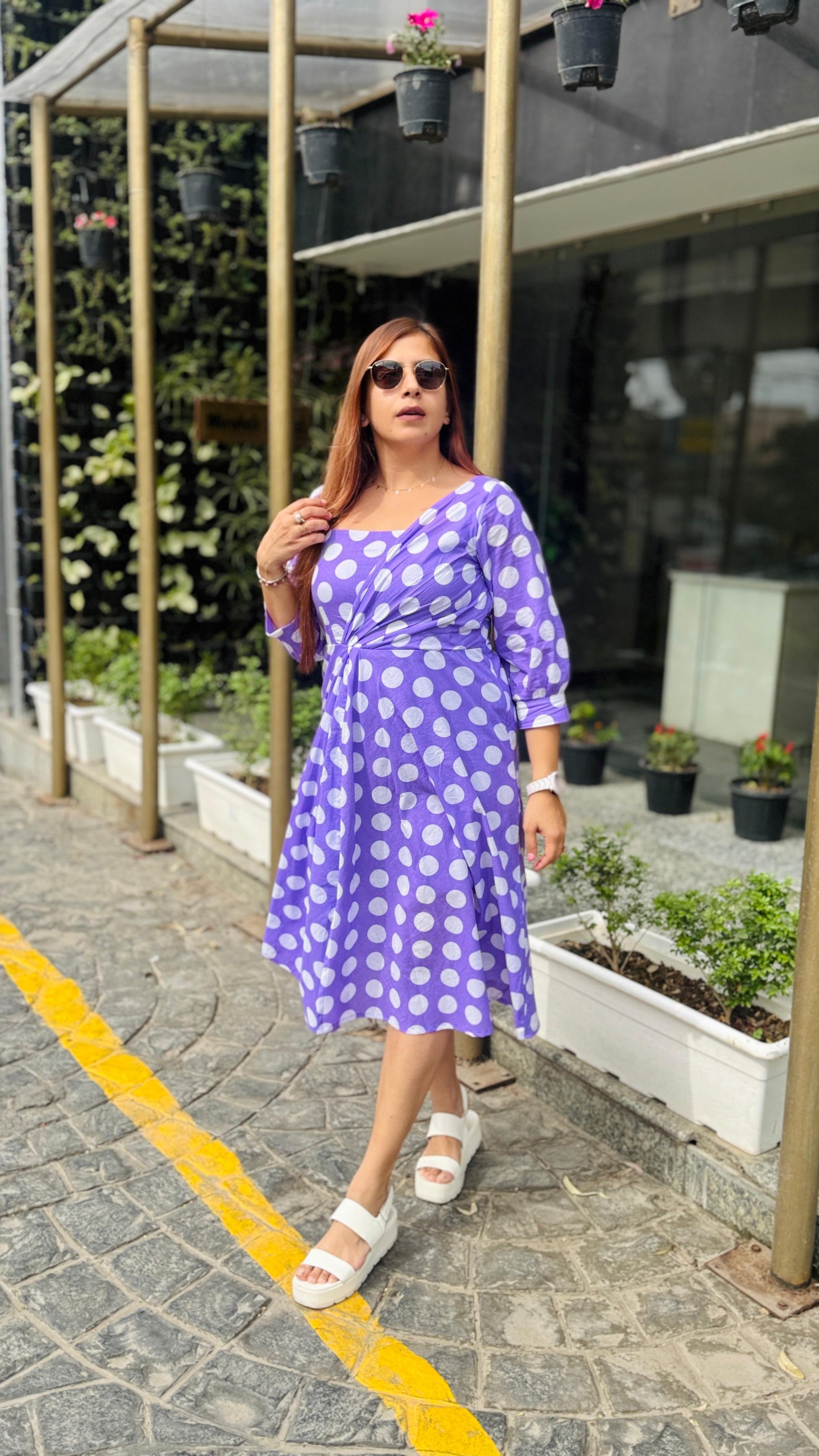 Woman in a purple polka dot dress standing outdoors near a building with plants.