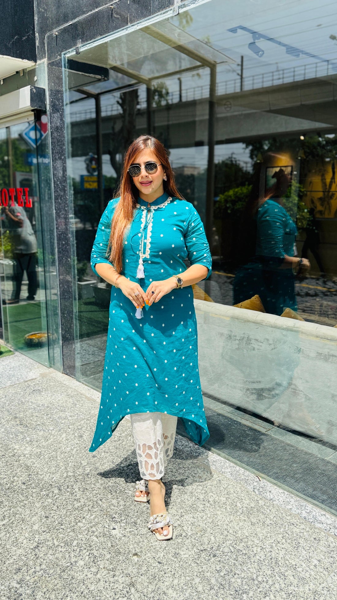 Woman in a teal kurta with white polka dots standing in front of a glass building.