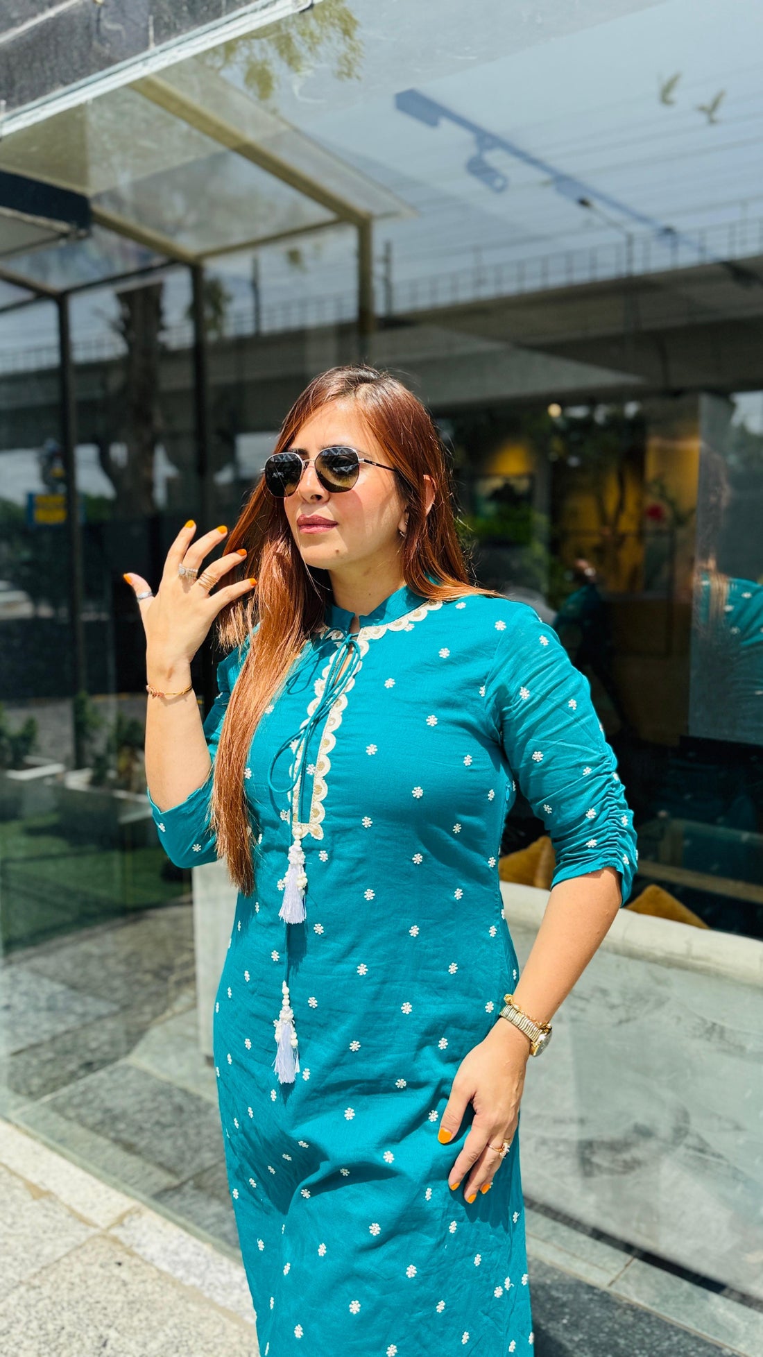 Woman in a teal kurta with white polka dots standing in front of a glass building.