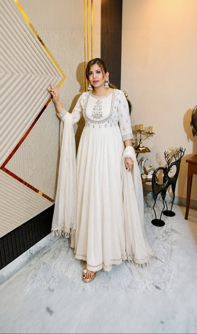 Woman in a white embroidered dress standing in an elegant interior setting.