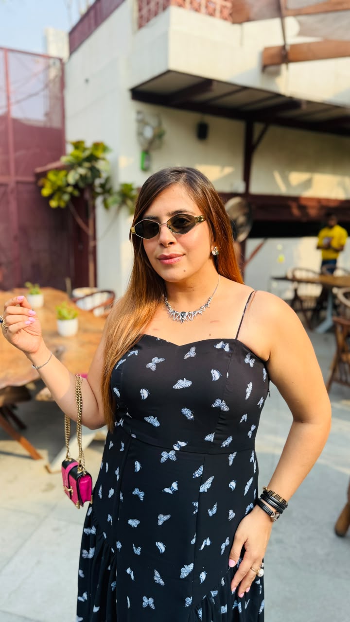 Woman wearing a black dress with white patterns, sunglasses, and a necklace outdoors.