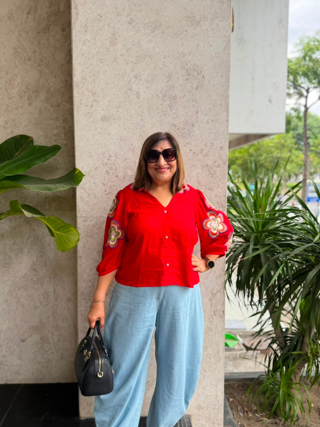 A woman wearing a bright red button-up blouse with gold embroidered details on the sleeves, paired with light blue wide-leg pants and white sneakers, standing in front of a modern concrete pillar with potted plants.