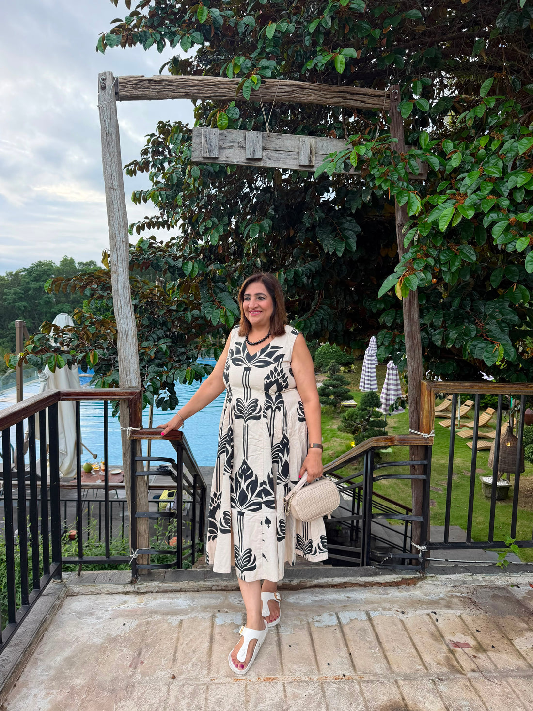 A woman posing on an outdoor balcony wearing a sleeveless beige dress with a black botanical pattern, holding a beige handbag and wearing white sandals, with a lush garden and pool in the background.