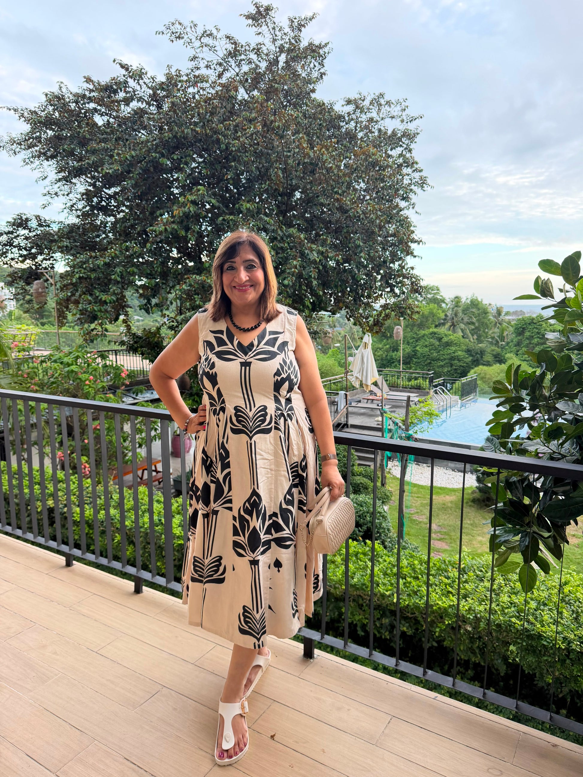 A woman posing on an outdoor balcony wearing a sleeveless beige dress with a black botanical pattern, holding a beige handbag and wearing white sandals, with a lush garden and pool in the background.