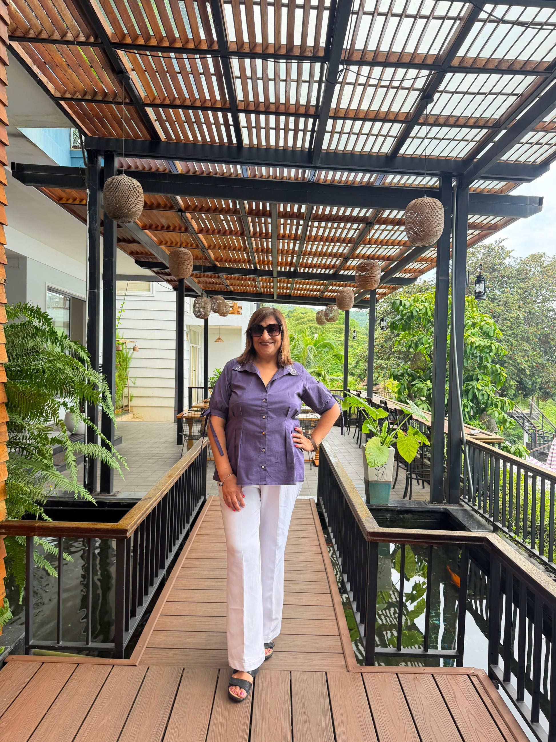 A woman wearing a purple short-sleeve button-up shirt and white pants stands on a wooden deck under a slatted roof structure with hanging woven baskets.
