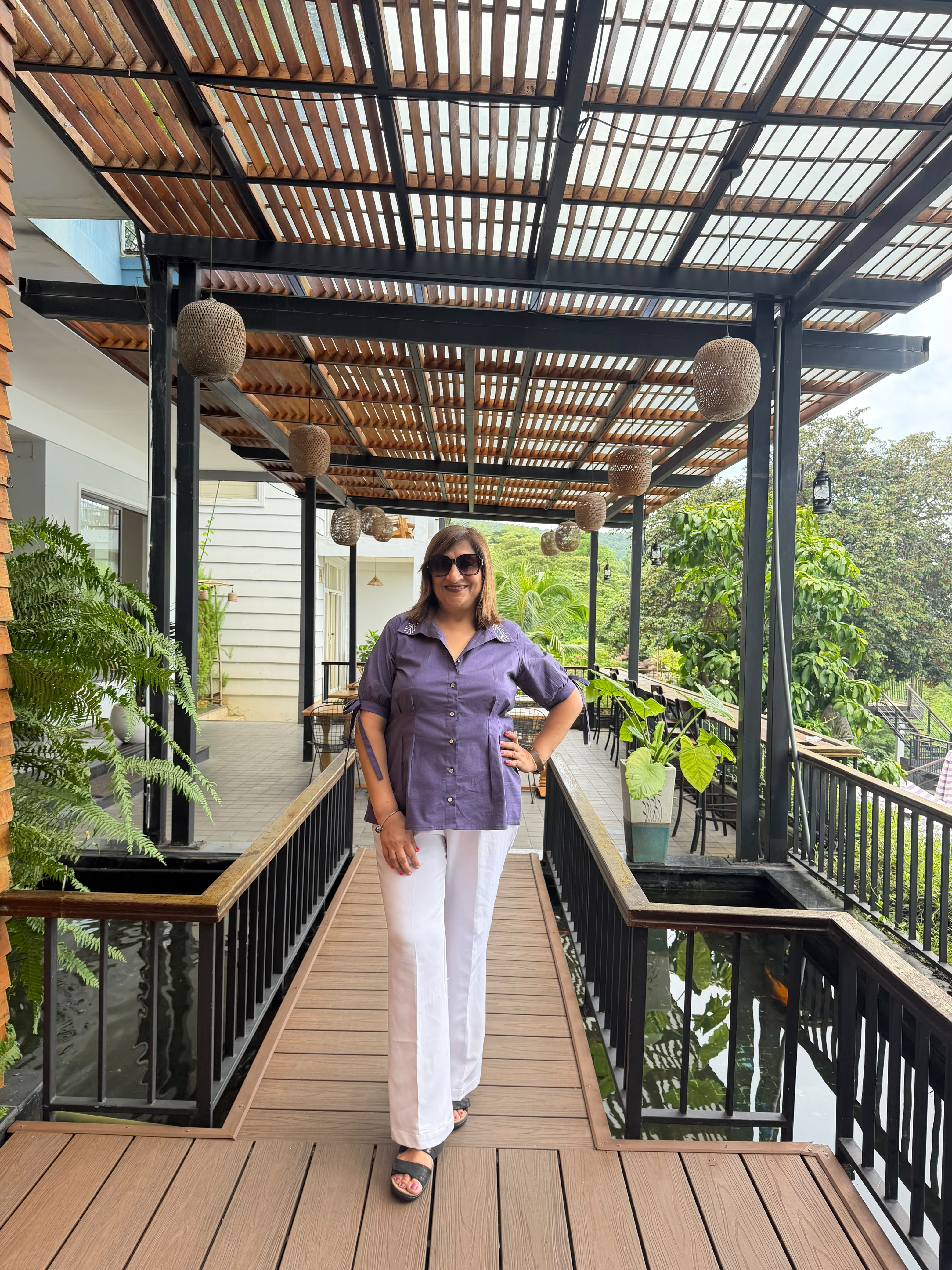 A woman wearing a purple short-sleeve button-up shirt and white pants stands on a wooden deck under a slatted roof structure with hanging woven baskets.