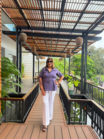 A woman wearing a purple short-sleeve button-up shirt and white pants stands on a wooden deck under a slatted roof structure with hanging woven baskets.