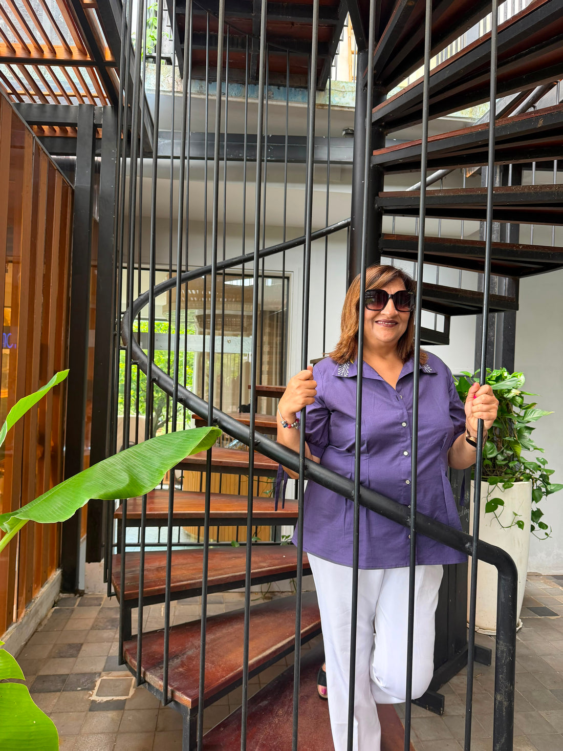 A woman wearing a purple short-sleeve button-up shirt and white pants stands on a wooden deck under a slatted roof structure with hanging woven baskets.