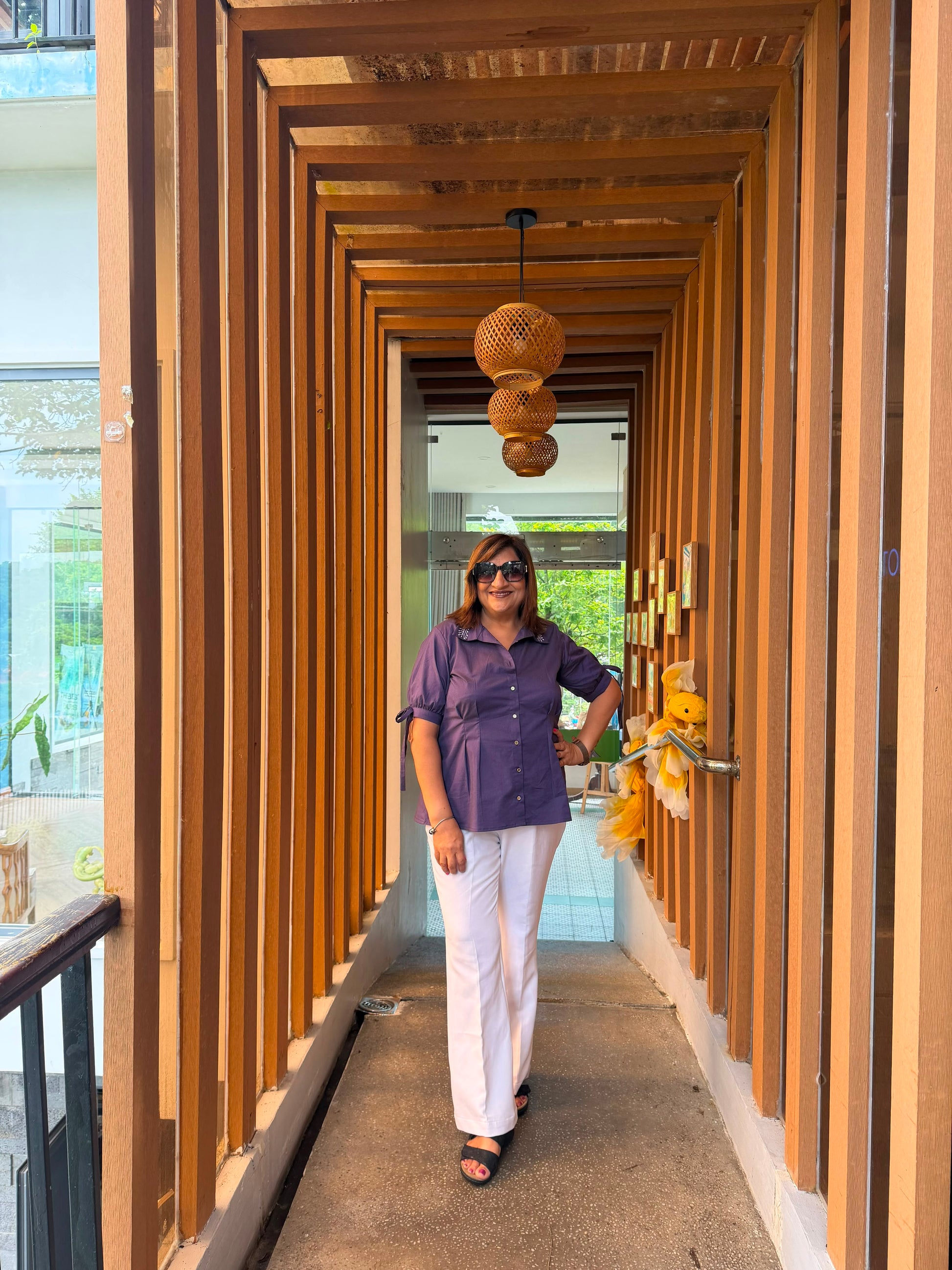 A woman wearing a purple short-sleeve button-up shirt and white pants walks down a modern wooden corridor with hanging woven pendant lights.