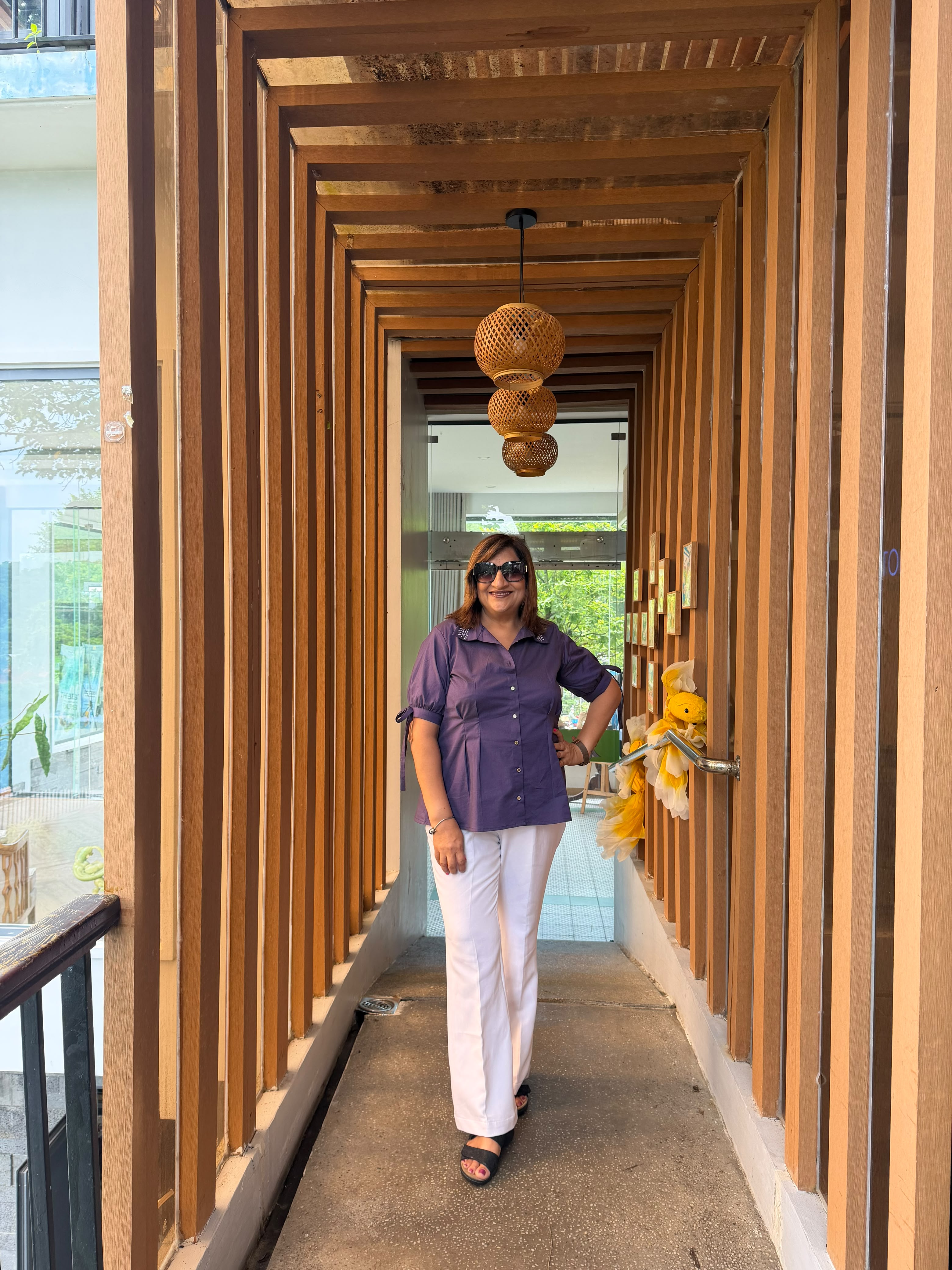 A woman wearing a purple short-sleeve button-up shirt and white pants walks down a modern wooden corridor with hanging woven pendant lights.