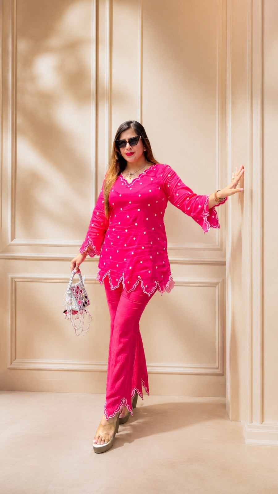 A woman modeling a bright pink traditional outfit consisting of a long-sleeve kurta and matching pants, accessorized with sunglasses, a small patterned handbag, and platform sandals, posing against a beige paneled wall.