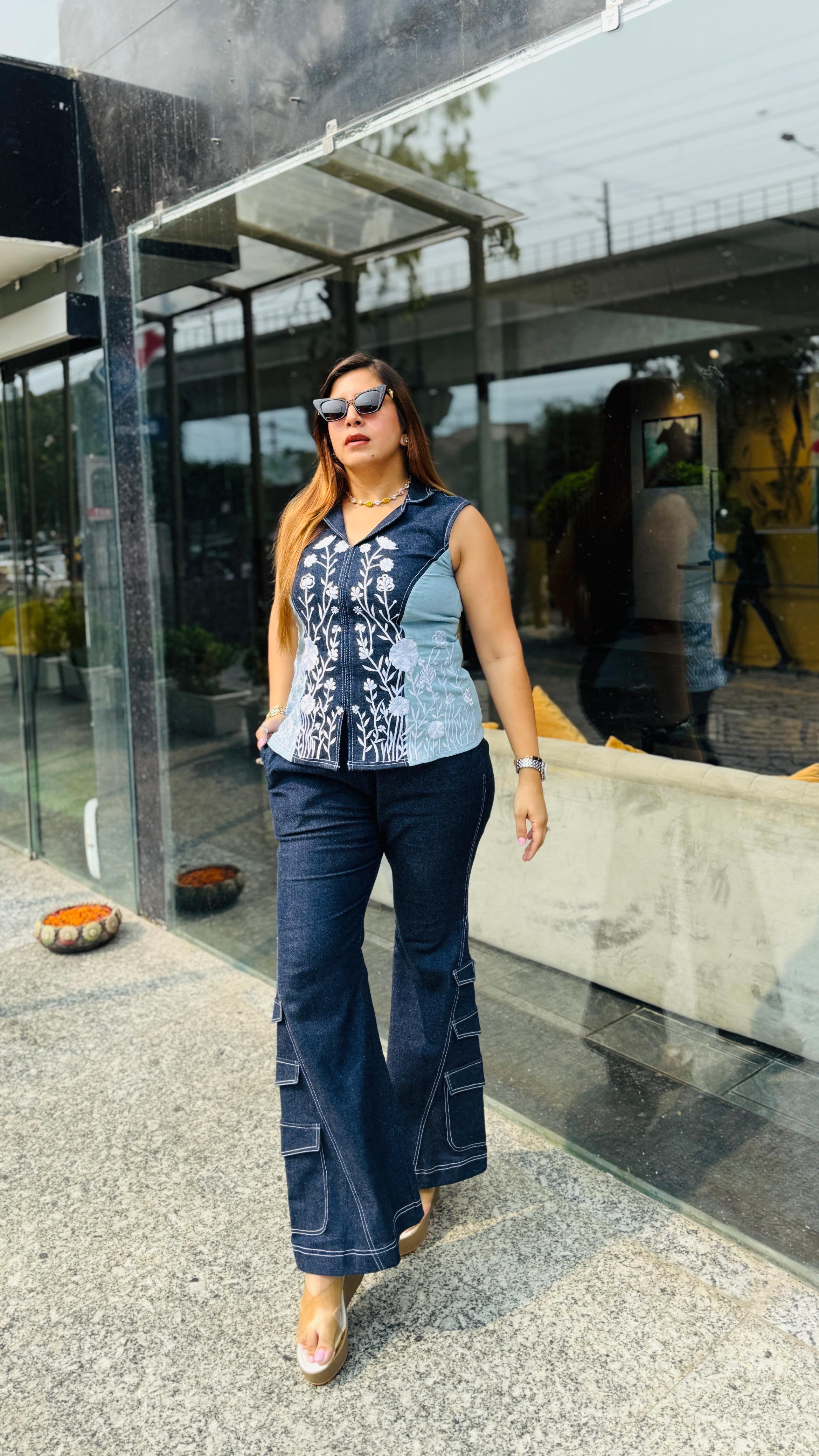 A woman walking outdoors in front of a modern glass building, wearing a sleeveless top with a blue and white patterned yoke and dark blue flared pants.